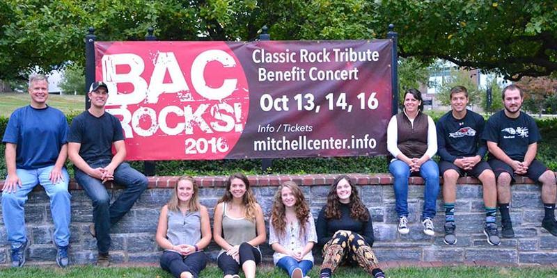A professor, students, and alumni participating in the BAC College Rocks concert pose in front of the banner at the entrance to campus