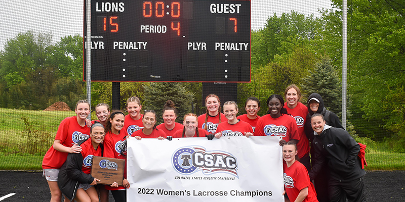 New champions women's lacrosse team stands in front of scoreboard.