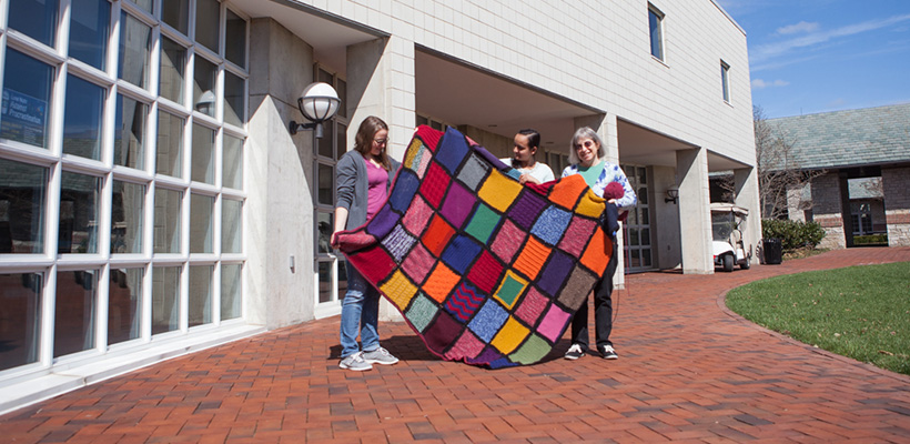 Bryn Athyn College library staff holding up a colorful afghan outside on the Brickman Terracebaskets full of knitting materials
